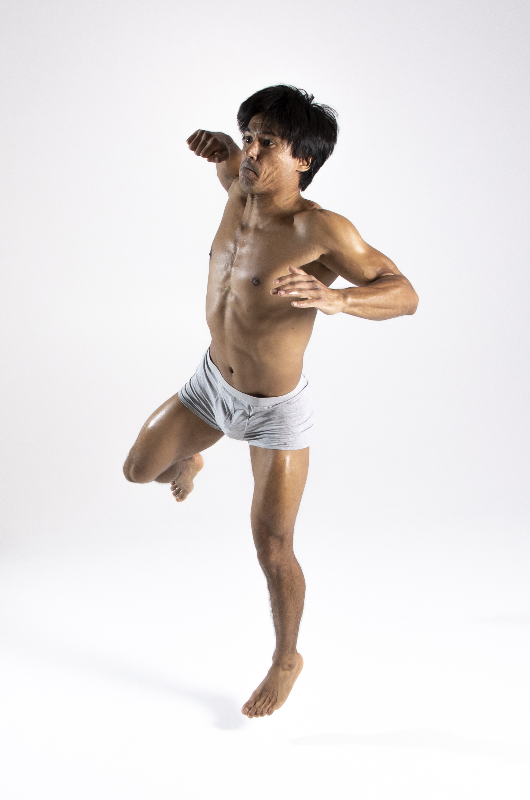 a shirtless asian man wearing grey underwear is standing barefoot in a fighting pose against a white background. he is muscular and fit, with one leg raised and arms positioned as if striking or blocking. the image appears to be a studio shot capturing a martial arts or action scene.