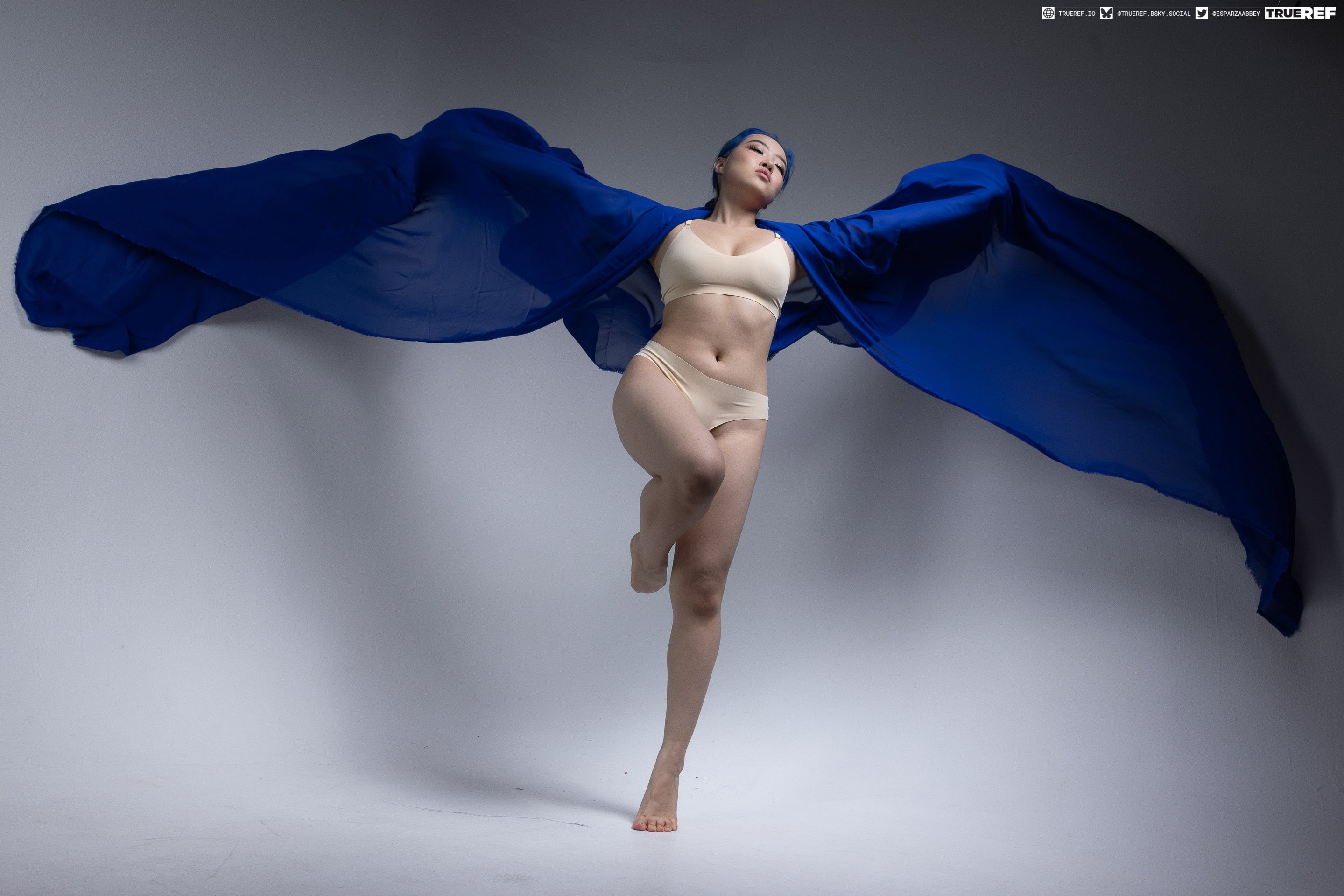 a full shot of an asian woman posing in a studio. she is barefoot and wearing a bra and panties set. a large, blue scarf is draped around her, creating a flowing effect. she is standing on one leg with the other knee raised, looking upwards.