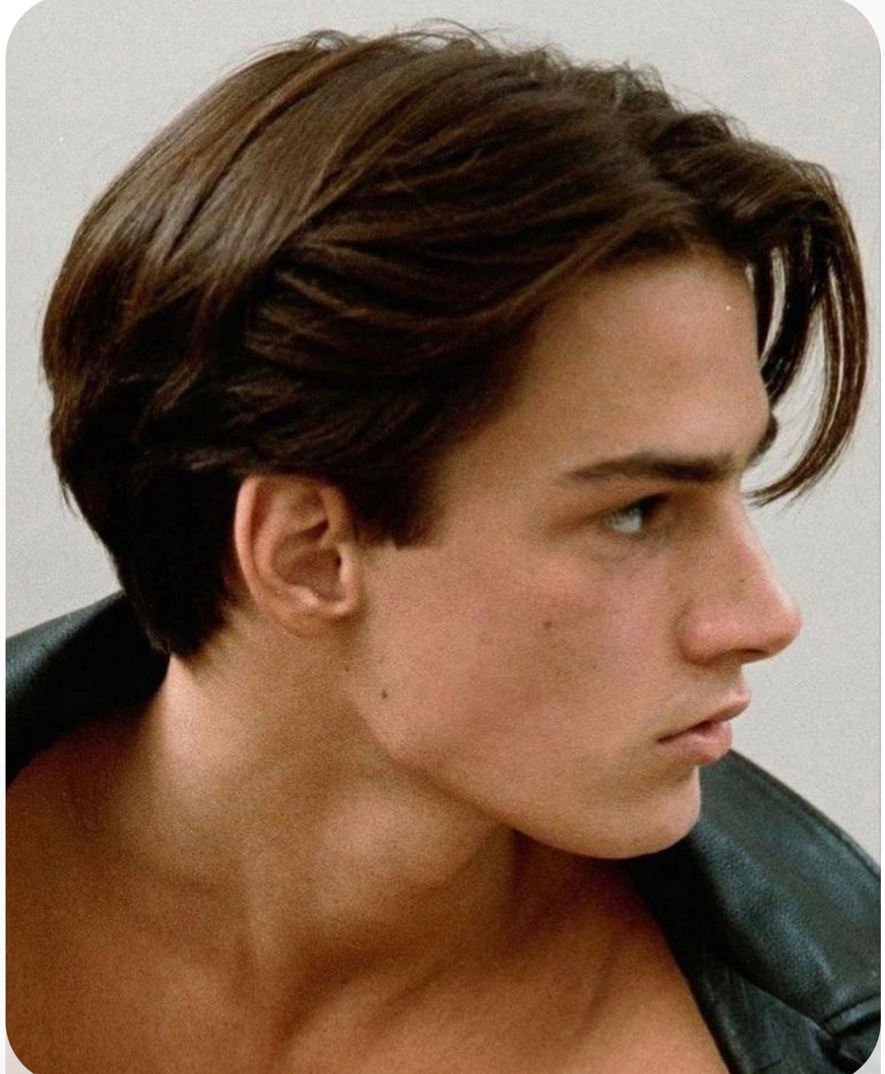 the image is a closeup portrait of a young man with a stylish haircut, taken from a slightly low and side perspective. he has brown hair styled with a center part and a slightly messy, textured look. the man has a neutral expression and is looking off to the right of the frame. he has a light complexion and is wearing a black leather jacket. the background is a light, neutral color and is blurred, keeping the focus on the man's face and hair. the lighting is soft and naturallooking, creating a flattering and approachable feel.