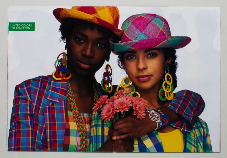 two women are posing for a united colors of benetton advertisement. they are wearing brightly colored plaid clothing and hats, large earrings, and holding flowers. the woman on the right has a colorful beaded bracelet. the overall style is reminiscent of 1980s or 1990s fashion.