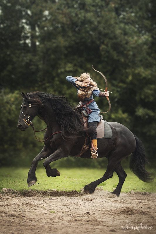 a woman riding a black horse through a forest, aiming her bow and arrow, in a historical recreation setting. she is dressed in a medievalstyle costume, with fur, leather and a blue tunic. the horse is a friesian. the image is wellcomposed, with the woman and horse in focus, and the forest out of focus in the background. it is a powerful image of a woman in the middle of a historic action.