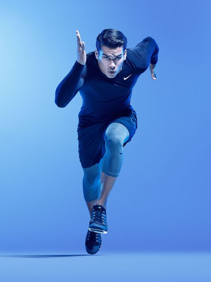 a man in a black nike running outfit is running, mid stride, against a bright blue background. he is looking directly at the camera with a determined look on his face. his arms are bent at the elbows and his hands are clasped in front of his chest. he is wearing black nike running shoes and his legs are extended as he runs. his expression is focused and determined. the photo captures the energy and intensity of running.