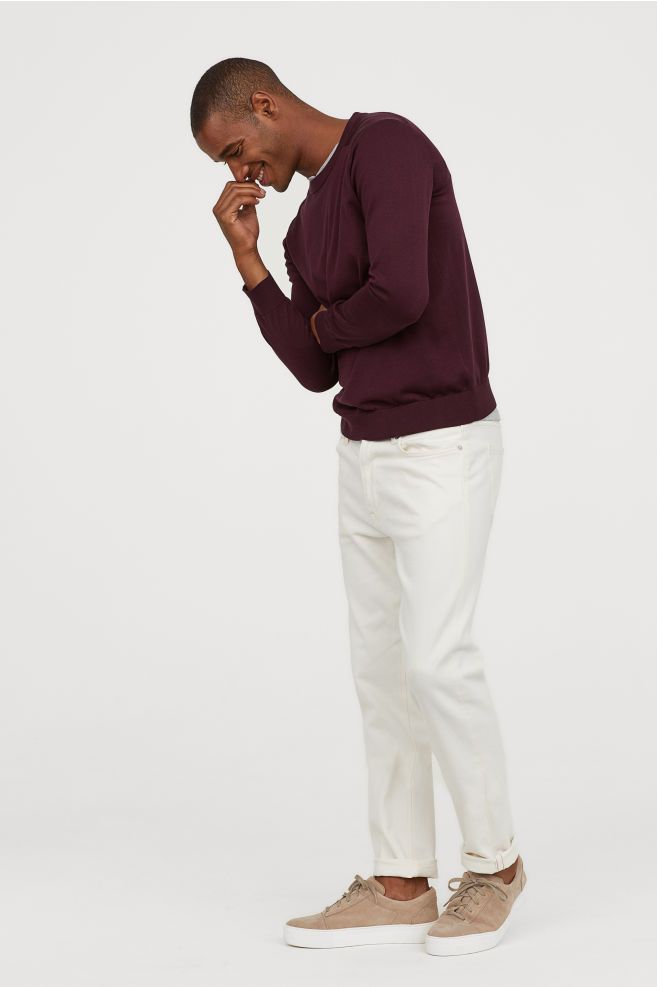 a man in a maroon sweater and white pants smiles as he looks down. he is wearing tan sneakers. the photo is taken against a white background.