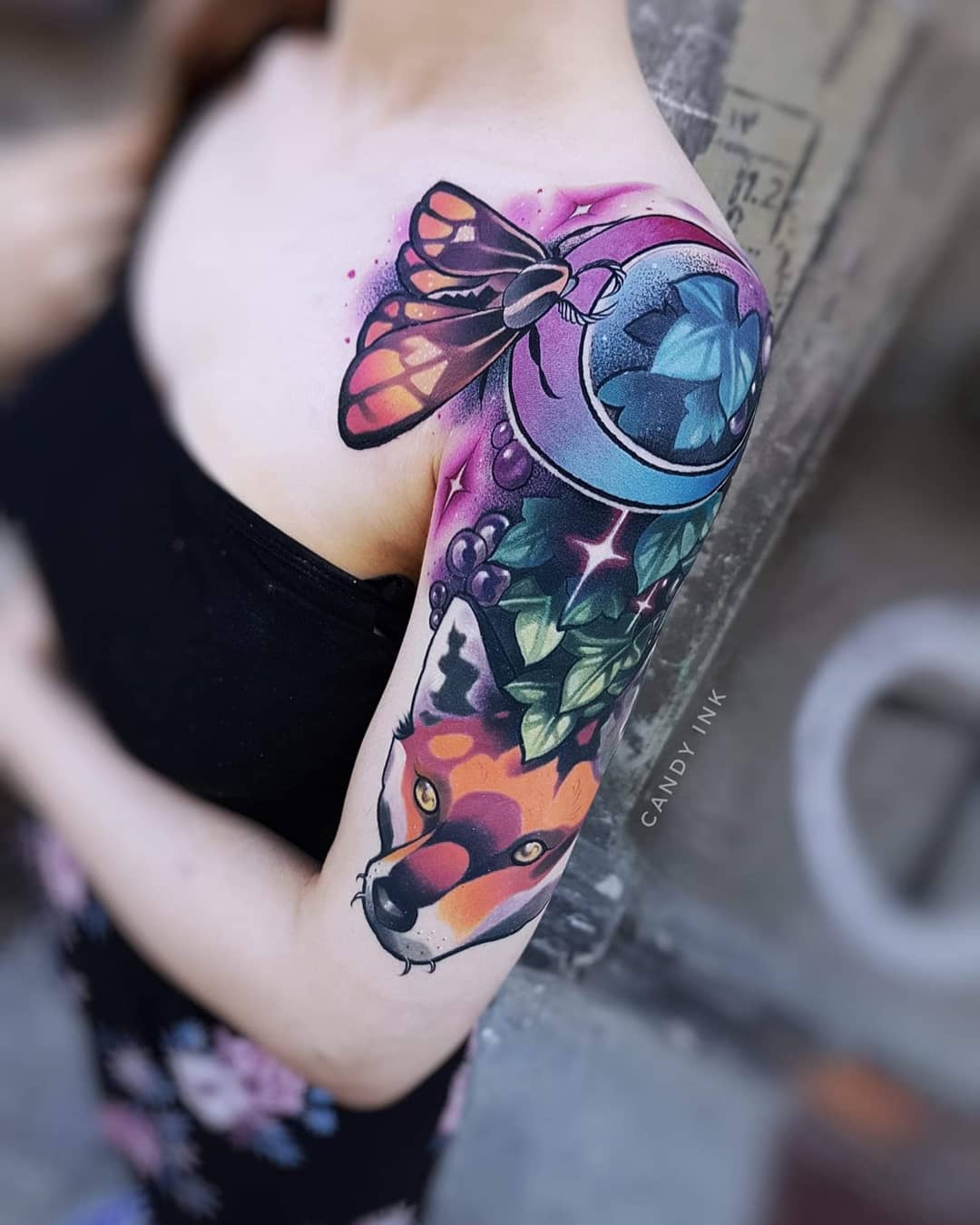 a closeup shot showcases a vibrant tattoo on a woman's left shoulder. the tattoo features a captivating combination of natureinspired elements.  a brightly colored butterfly with orange and black wings perches atop a stylized blue and purple moon adorned with delicate green leaves.  trailing down the arm are lush green grapevines laden with plump purple grapes, interspersed with sparkling white stars. emerging from beneath the vines is the head of a majestic fox with piercing orange eyes and a captivating gaze. the tattoo's artistry is evident in the intricate details and the harmonious blend of colors. the background provides a glimpse of the tattoo parlor setting.