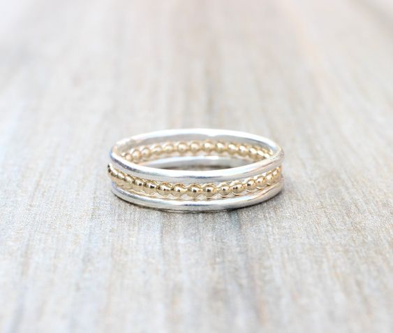 a set of three stacking rings lay on a light gray wooden surface. the top and bottom rings are silver, while the center ring is gold. all three rings feature small beaded details along the band.