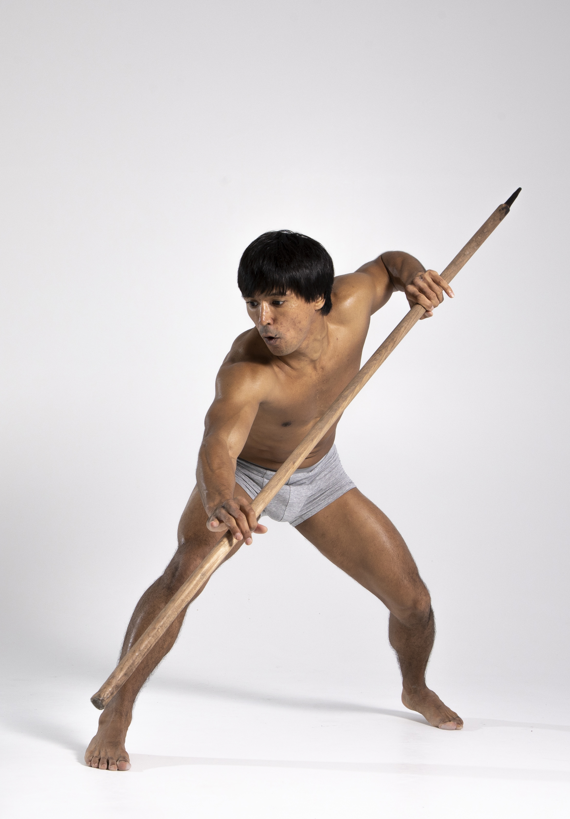 a barechested man with dark hair is in a dynamic pose, holding a wooden spear. he appears to be a warrior or fighter, clad in a loincloth. the studio shot captures his muscular physique against a plain white background.