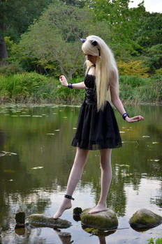 a young blonde girl in a black gothic lolitastyle dress stands in a calm pond, stepping on rocks.  she has long blonde hair and is wearing black jewelry. the setting is a peaceful forest or woodland area with lush greenery and trees reflected in the water. the overall mood is whimsical and slightly ethereal.