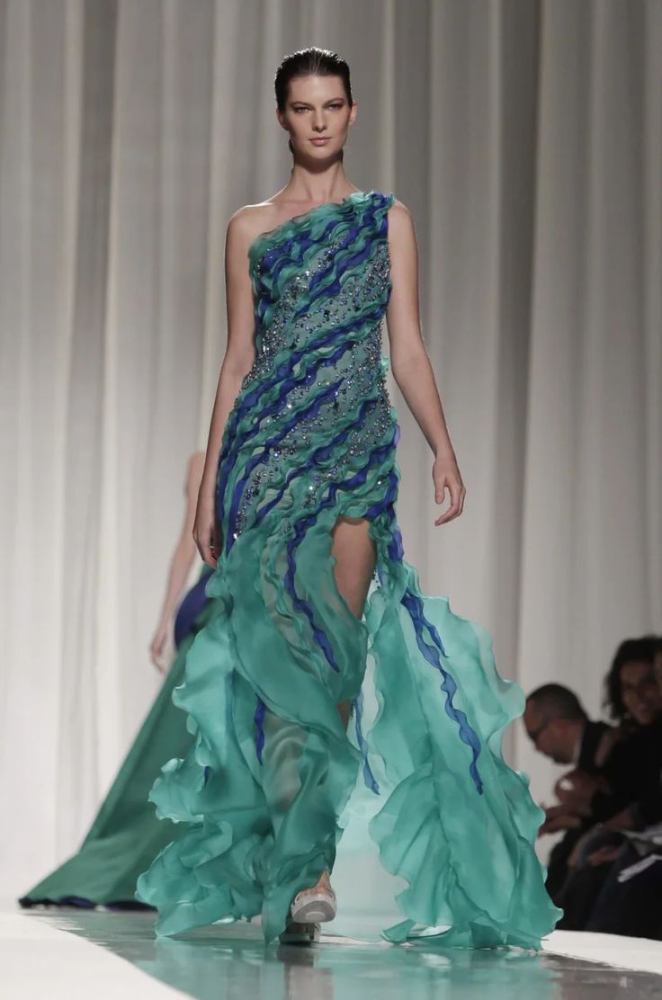 a beautiful model in a blue and green dress with ruffles walks down the runway. the dress is very stylish and elegant. the model is posing for the cameras and her makeup is flawless. the background is a blurry backdrop of a fashion show.