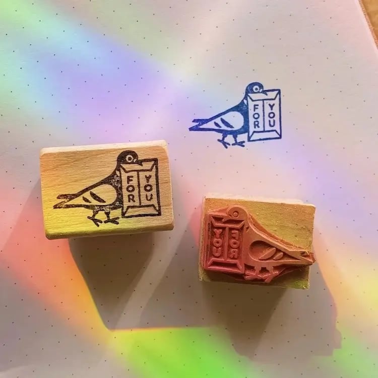 the image shows three small, wooden rubber stamps featuring a stylized pigeon carrying a package labeled "for you."  two of the stamps are lightcolored wood with dark ink, and one is a darker, reddishbrown wood with dark ink. the stamps are arranged on a speckled, offwhite surface. a rainbowlike light effect is visible across the background, adding a colorful, whimsical element. the overall impression is of cute, handmade stationery or craft supplies.