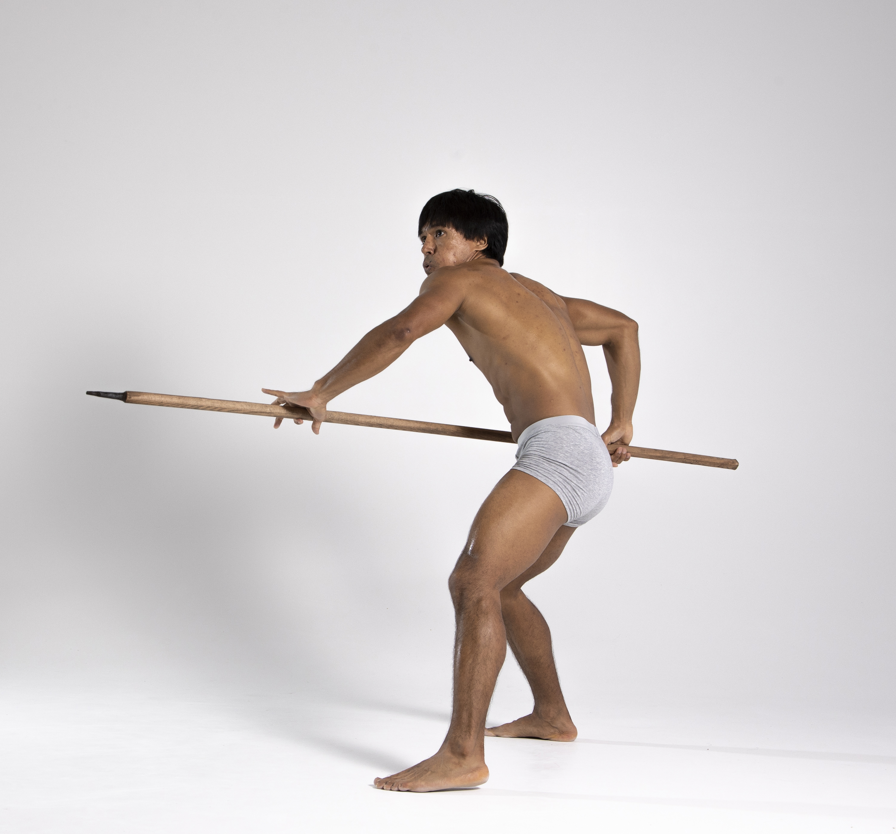 a man posing with a spear in a studio shot. he is barefoot and wearing grey underwear. he has dark hair and is muscular. the background is white.