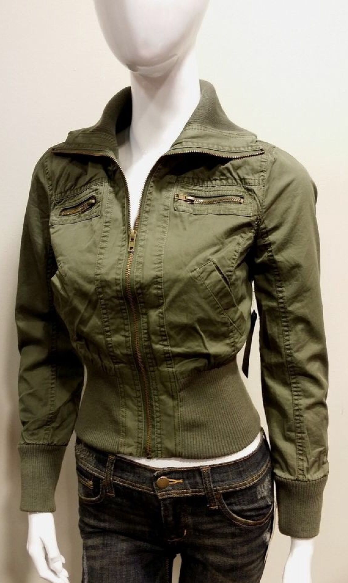 a mannequin is wearing a green jacket with a zipper closure. the jacket has pockets on the chest and a ribbed waist band. the mannequin is also wearing jeans. the photo is taken from the front and shows the full body of the mannequin.