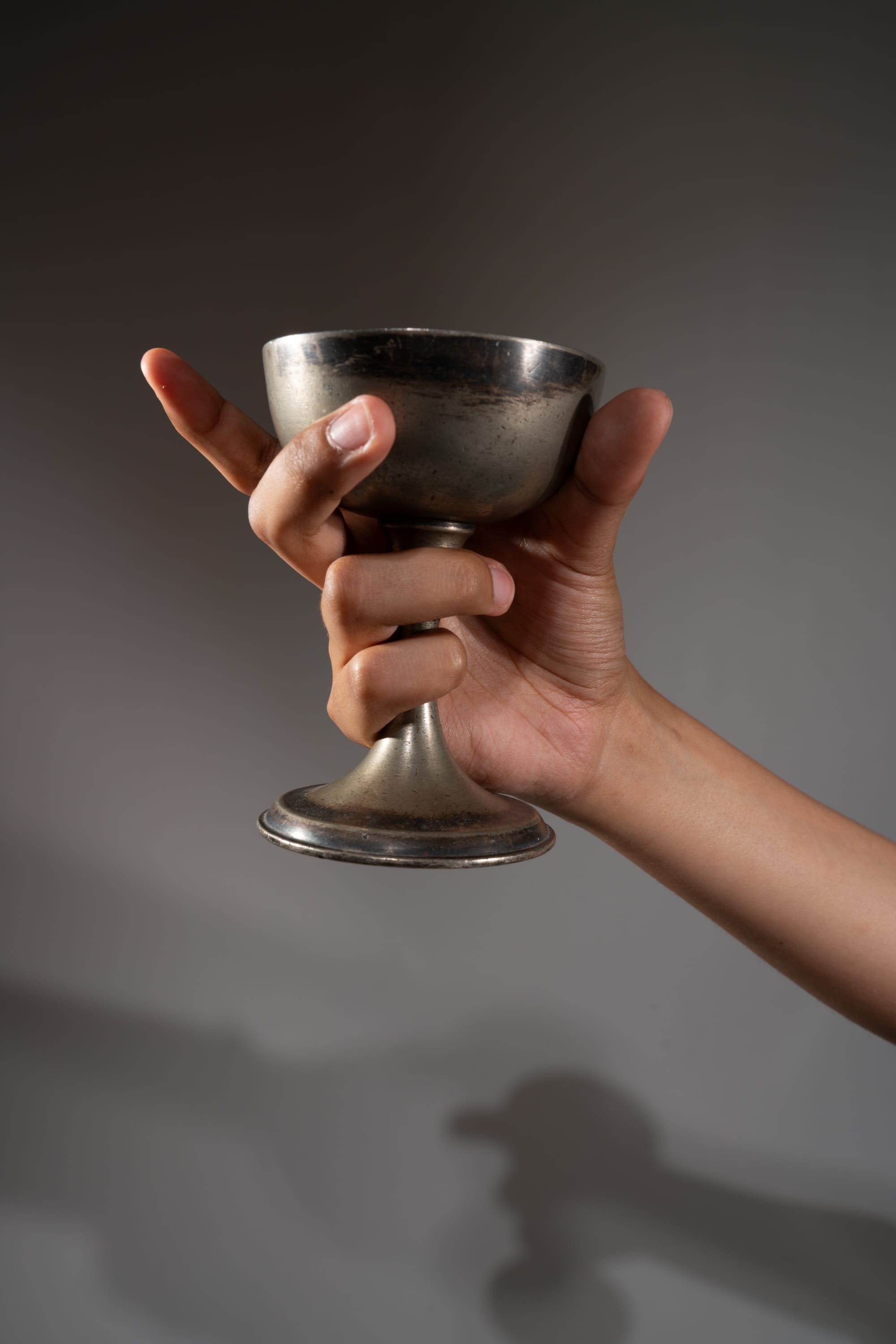 a hand is holding a silver, metal goblet up in the air in front of a gray background.
