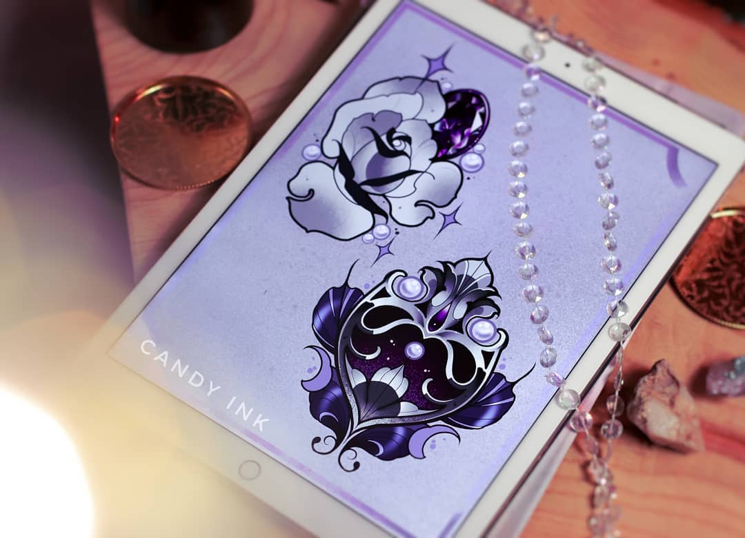 the image shows a tablet showcasing two tattoo designs on a purple background. the designs feature a white rose with a purple gem and an ornamental heart with a purple gem and cosmic elements. they are drawn in a neotraditional style. the tablet is surrounded by crystals, beads, and a coin.  the scene is lit with warm, artificial light.
