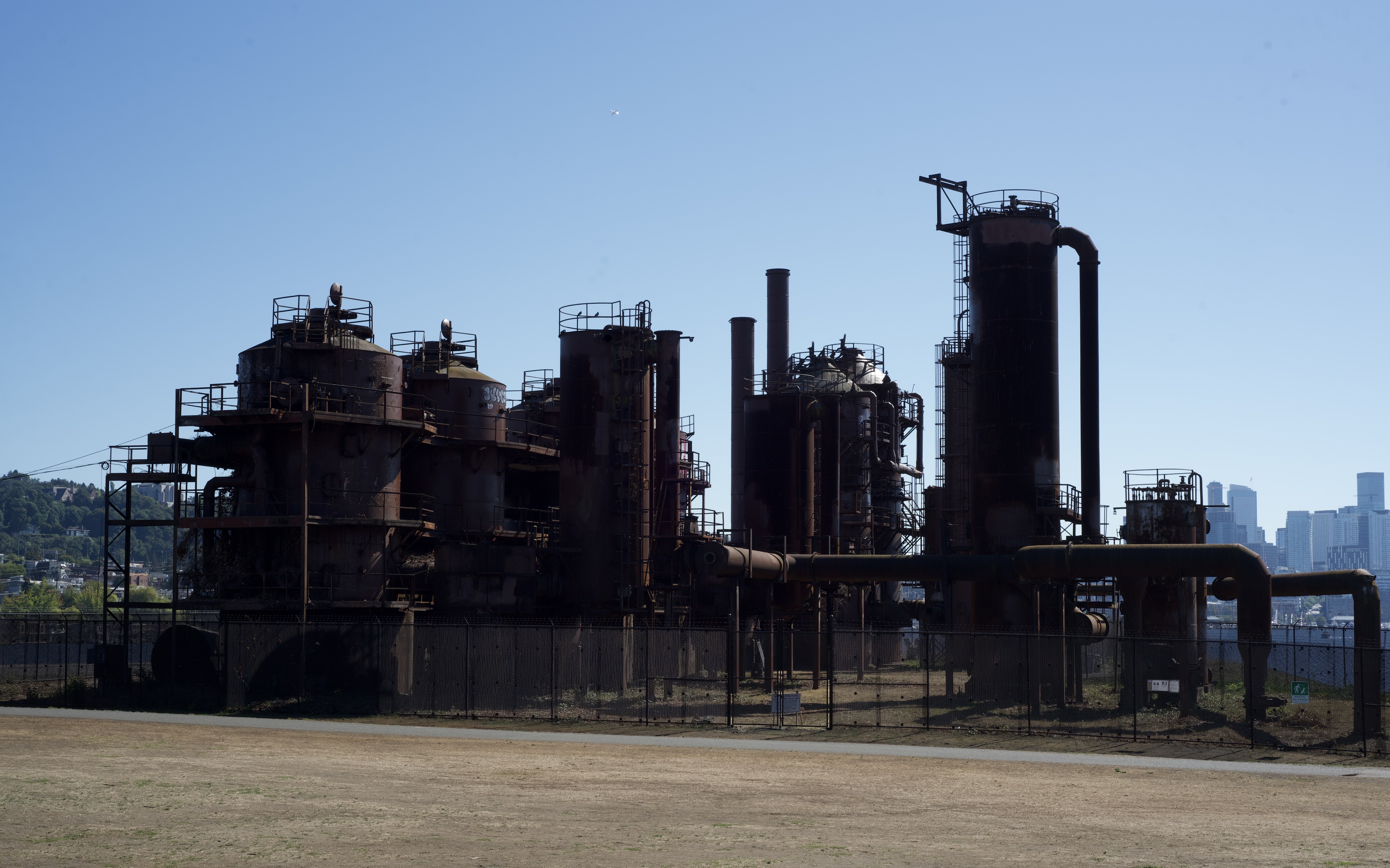 a full shot of the gas works park in seattle, washington, on a sunny day. the foreground shows a grassy area and a black fence surrounding the rusty industrial structures. in the background, a portion of the seattle skyline is visible. the image showcases the contrast between the aged industrial structures and the modern city. the structures are dark silhouettes against the bright sky.