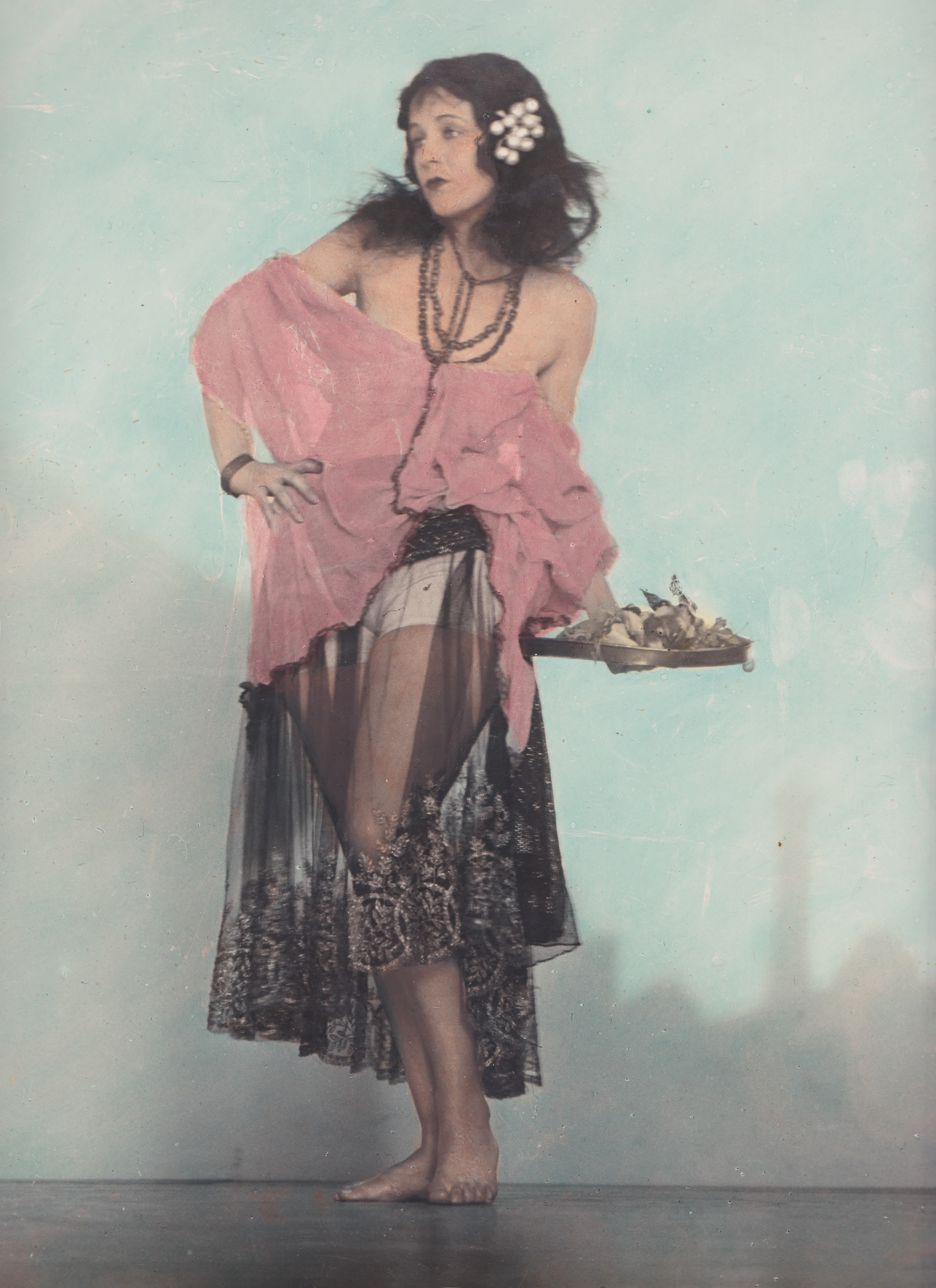 the image is a vintage, handcolored photograph of a young woman, possibly a dancer, from the 1920s or 1930s. she is elegantly dressed in a sheer black lace dress over pink undergarments, with a pink sheer shawl draped over her shoulders.  she wears beads around her neck and has flowers in her hair.  she's holding a small tray with what seems to be food or offerings.  the background is a plain, light teal backdrop common in studio portraits of the era. the overall style suggests an art deco influence.  the woman has a slightly surprised or curious expression on her face. the photograph has a soft focus and the colors are slightly muted, characteristic of the handcoloring process.