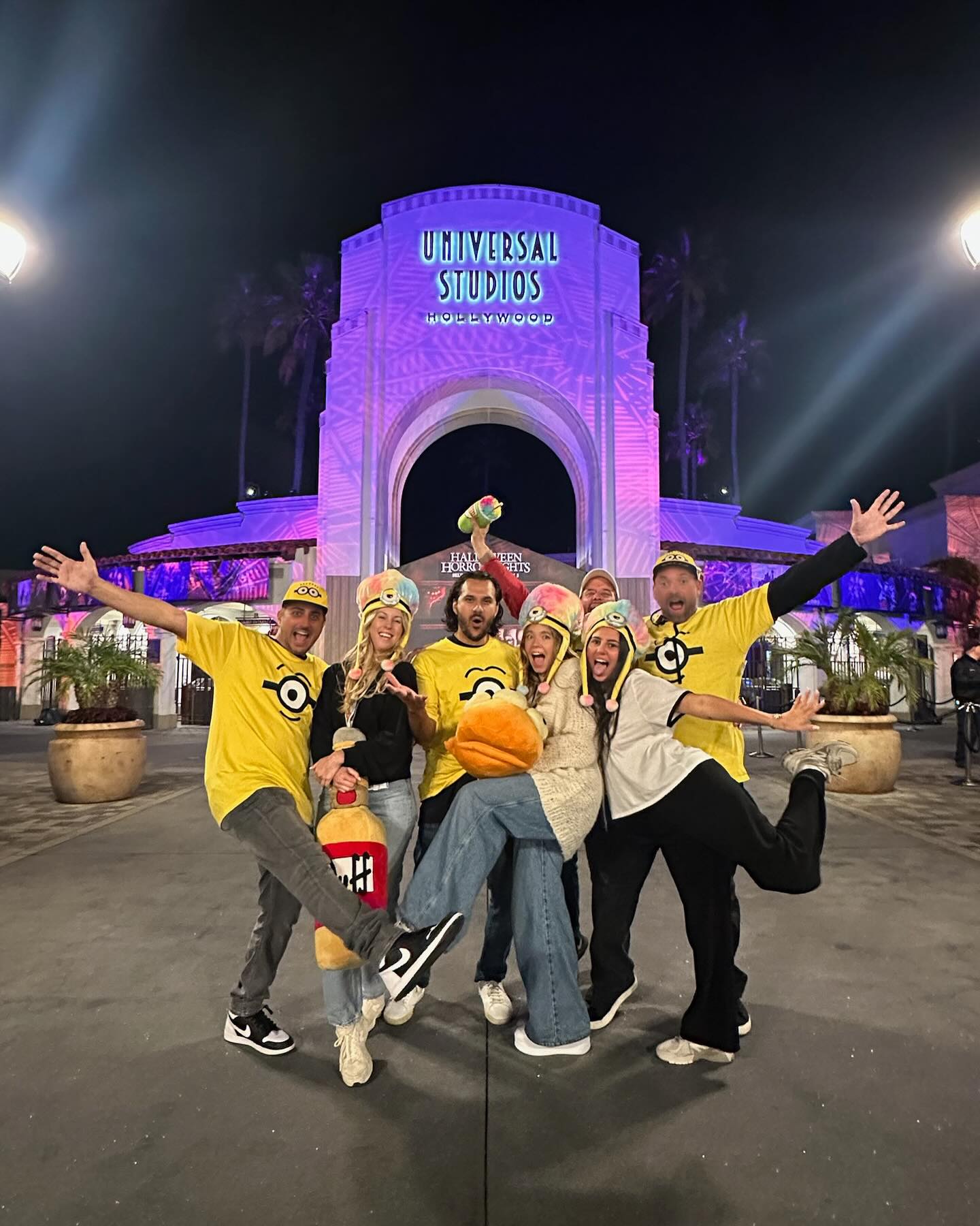 a group of friends, dressed in minionthemed costumes and hats, pose for a photo in front of the iconic universal studios hollywood archway at night.  the archway is illuminated in purple, signifying the halloween horror nights event. the friends appear joyful and excited, capturing a memorable moment during their visit to the theme park.  the photo captures the energy of the event and the overall celebratory atmosphere.