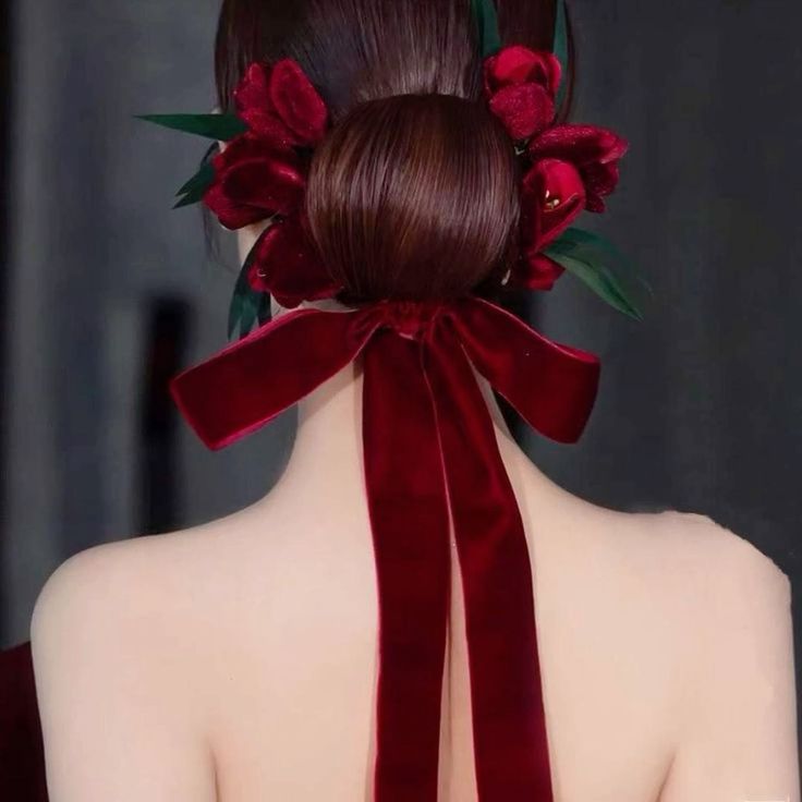 the image shows the back of a woman's head featuring a meticulously styled dark brown bun adorned with a cluster of dark red velvet flowers and a long, elegant dark red velvet ribbon tied in a bow. the woman's bare shoulders and back are visible, and the overall style is elegant and sophisticated, suggestive of a wedding or formal event.  the backdrop is a simple, dark gray, which contrasts beautifully with the rich colors of the hair accessory.