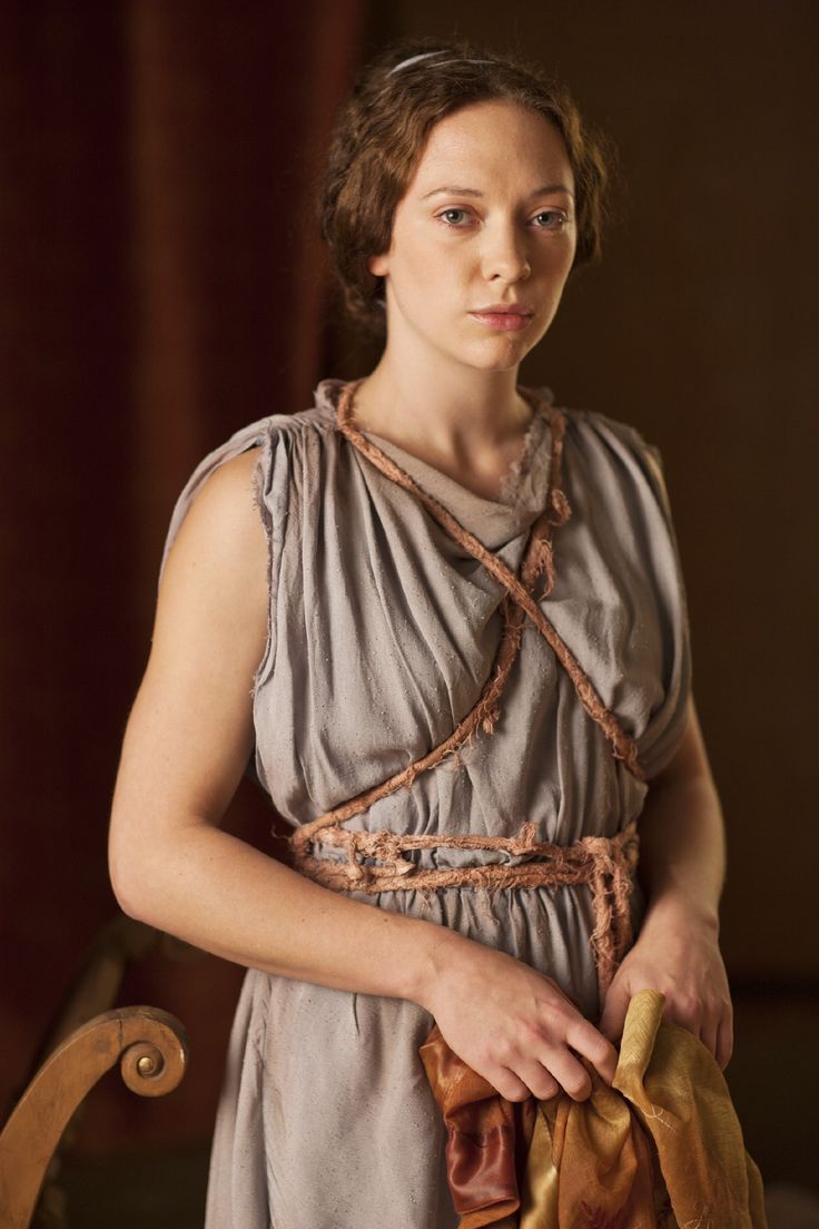 a closeup portrait of a woman, possibly an actress in a historical drama, dressed in a simple, light grey garment resembling an ancient greek or romanstyle dress. the dress is loosely fitted and cinched at the waist with a ropelike belt.  the fabric has a slightly frayed texture. the woman's hair is styled in a wavy updo, and she wears a thin headband. she has a serious, almost pensive expression, and she gazes directly at the camera.  she's holding a folded piece of fabric in her hands, which appears to be a different material and color than her dress. the background is blurry, suggesting a softly lit studio setting with a dark, possibly burgundy, curtain visible. a portion of a wooden chair arm is visible in the bottom left corner. the overall mood is somber and contemplative.