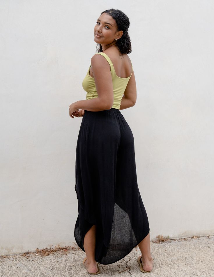 a young woman with curly hair is standing in front of a white wall. she is wearing a lime green tank top and black wideleg pants. the pants have a unique draped and asymmetrical design. she is posing for the camera with a slight turn, giving a view of the back of her outfit. the background is a simple white wall with a bit of ground visible at the bottom. the overall image has a bright and airy feel due to the natural lighting.