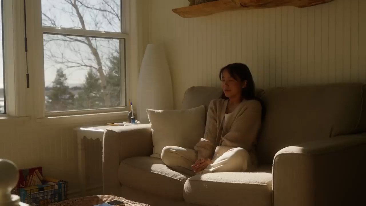 a woman sits on a beige couch in a sundrenched living room, bathed in the warm glow of natural light streaming in from a large window. the room is bright and airy, with a peaceful and serene atmosphere. the woman is dressed in comfortable, neutraltoned clothing, suggesting a sense of relaxation and contentment. the image evokes a feeling of quiet contemplation and the simple joys of being at home.