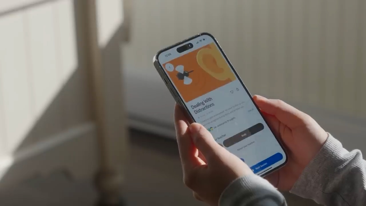 the image shows a closeup shot of a person holding a smartphone in their hands. the screen displays a mindfulness app, with a session titled "dealing with distractions" in focus. the app interface features a minimalist design with a calming color scheme. the image suggests a user engaged in a selfimprovement activity, likely in the comfort of their bedroom, as indicated by the surrounding environment and the soft natural lighting.
