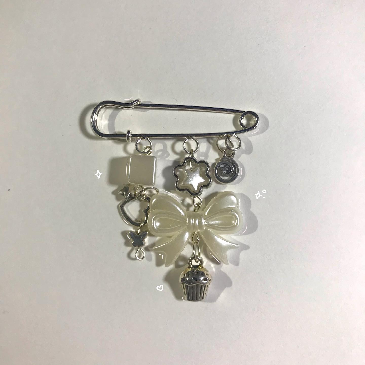 a closeup shot of a handmade brooch on a white background. the brooch is a large silver safety pin with several charms hanging from it, including a white bow, a silver cupcake, a silver flower, a silver star, and a white square. there is also a butterfly charm connected to the bow and the square charm. the background is plain white.