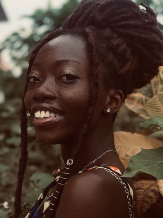 a closeup portrait of a young african woman with dark skin and long, dark dreadlocks styled in a high bun. she is smiling, and her expression is one of happiness and confidence.  she's wearing simple silver jewelry, a necklace and small earrings. the background is softly blurred, showing lush green foliage, creating a natural and relaxed setting. the image focuses on the woman's face and upper body, highlighting her features and her rich, dark skin tone. the overall style is warm and natural.