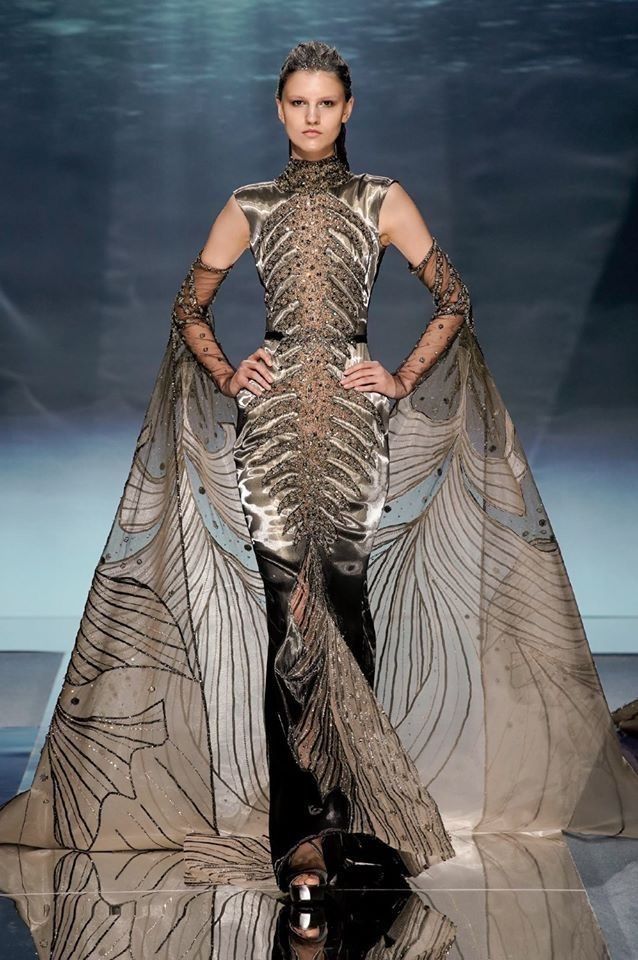 a woman is walking down a runway wearing a metallic gown with skeletonlike embellishments down the front. the gown has a high neck and long sleeves made of sheer fabric. the dress has a cape attachment with a winglike pattern. the model is posing with her hands on her hips. the background is a blurred blue color.