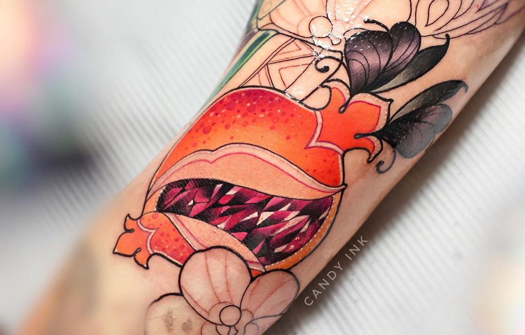 a closeup photograph showcasing a vibrant neotraditional tattoo of a pomegranate on an individual's forearm. the pomegranate is depicted with an open mouth, revealing rows of sharp teeth. the tattoo is rendered in a bold and colorful style, with shades of orange, red, and purple. surrounding the pomegranate are delicate floral elements, including a partially completed flower in the foreground. the tattoo artist's watermark, "candy ink," is subtly placed near the wrist. the image is brightly lit, highlighting the intricate details of the tattoo.