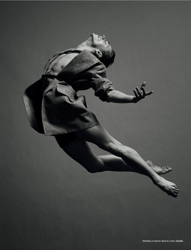 a black and white image of a man in a coat, midjump. he is facing forward with his body bent in a slight arc, with his head titled back. his legs are stretched out and his arms are outstretched, one of them is bent. the backdrop is a simple grey background.