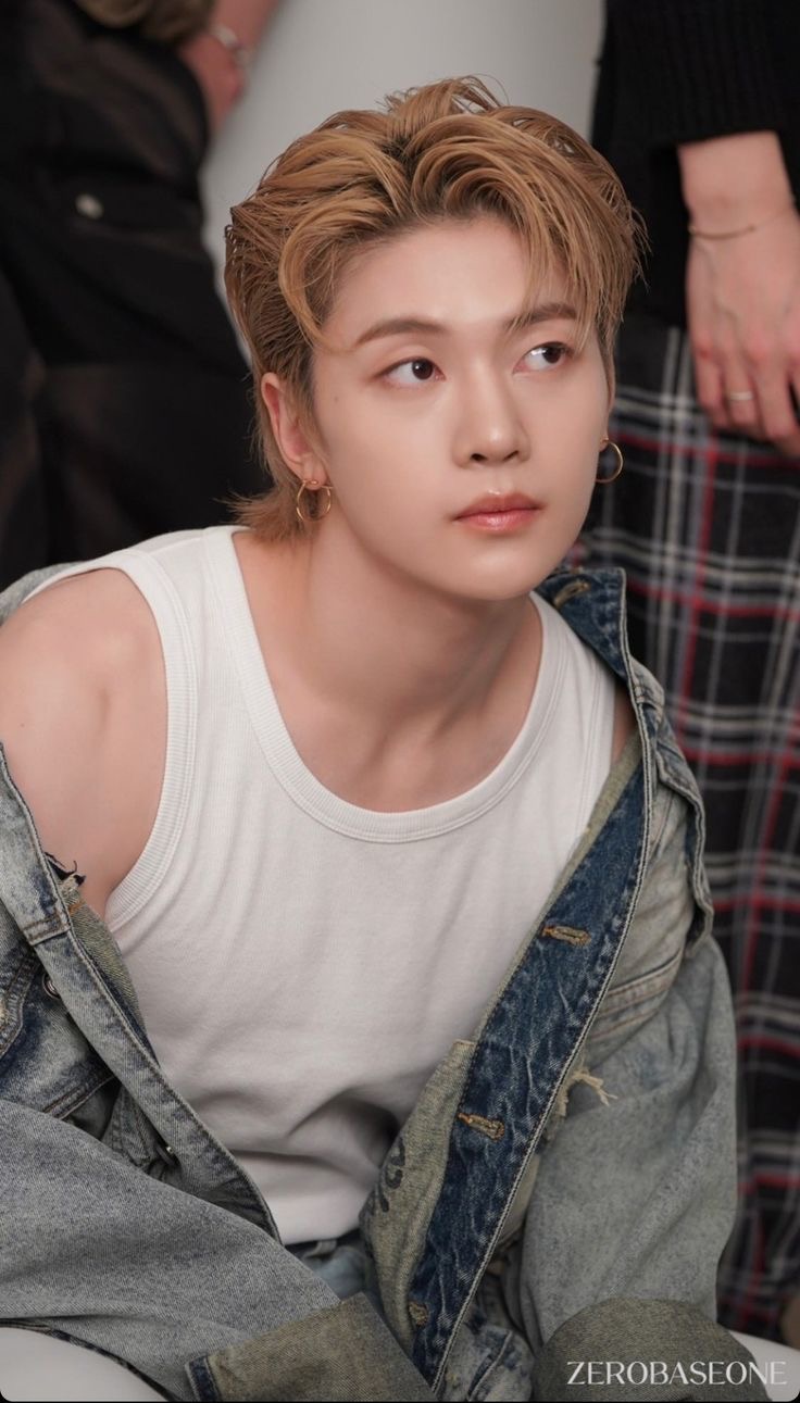 this is a portrait of kim jiwoong, a member of the kpop group zerobaseone. he is styled with blonde hair and is wearing a white tank top and an oversized denim jacket. he is also wearing a small hoop earring. the background features glimpses of other people and clothing, indicating this might be a photoshoot setting. the lighting is soft and natural, creating a warm and approachable feel to the image.
