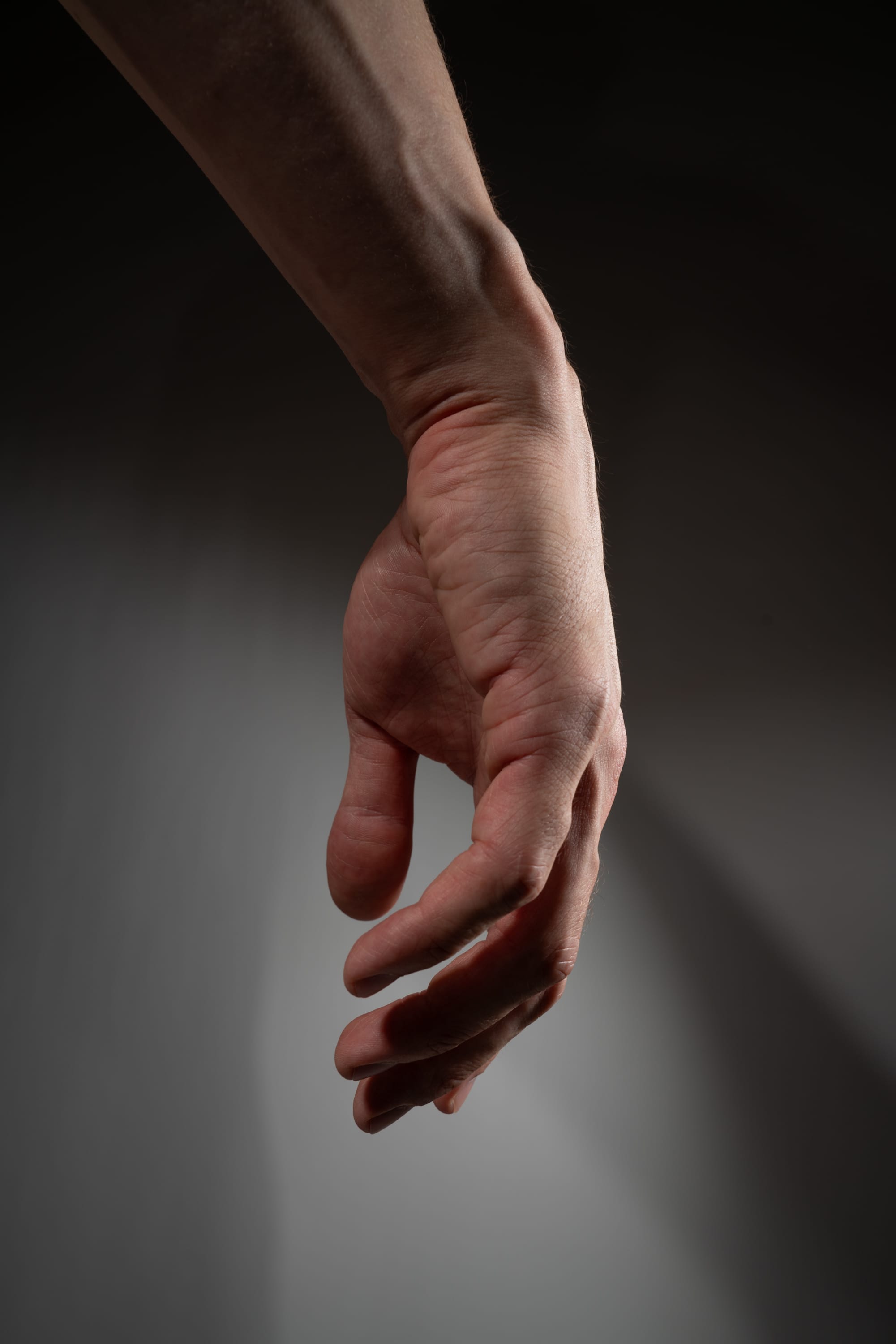 a man's right hand is extended downward against a gray background. the hand is relaxed, with the fingers slightly curled. the light source is coming from the left side, creating shadows on the right side of the hand. the image is a closeup, focusing on the details of the hand and arm.