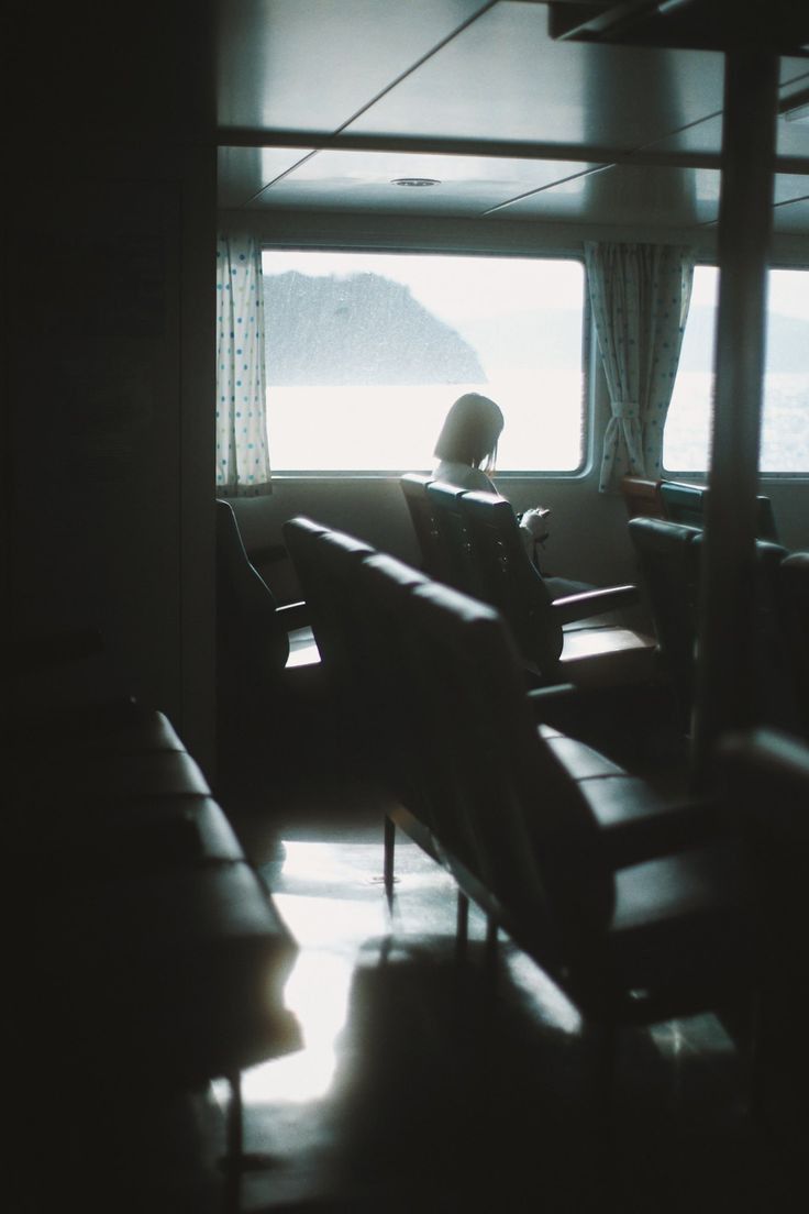 a woman sits alone on a ferry, looking out at a serene seascape. the interior of the ferry is dimly lit, with rows of dark leather seats. sunlight streams in from the large window, illuminating the woman's figure and casting shadows around the space. the scene conveys a sense of peace and solitude.