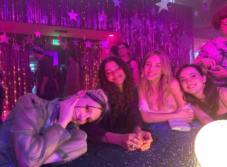 a behindthescenes photo of the cast of the hbo series euphoria at what appears to be a promthemed photoshoot.  zendaya, maude apatow, hunter schafer, and barbie ferreira are seated together at a table, smiling and looking happy. the setting is decorated with purple and pink lights and stars, creating a glamorous and celebratory atmosphere. the actresses are dressed in stylish attire suitable for a prom or party. the image evokes a sense of friendship and camaraderie among the cast members.