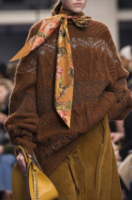 closeup shot of a fashion model showcasing a brown chunky knit sweater with a subtle pattern, paired with mustard yellow corduroy pants and a floral silk scarf tied around the neck.  the model also carries a mustard yellow handbag with a gold chain strap. the overall aesthetic is autumnal, warm, and sophisticated. the image is likely from a fashion runway show.