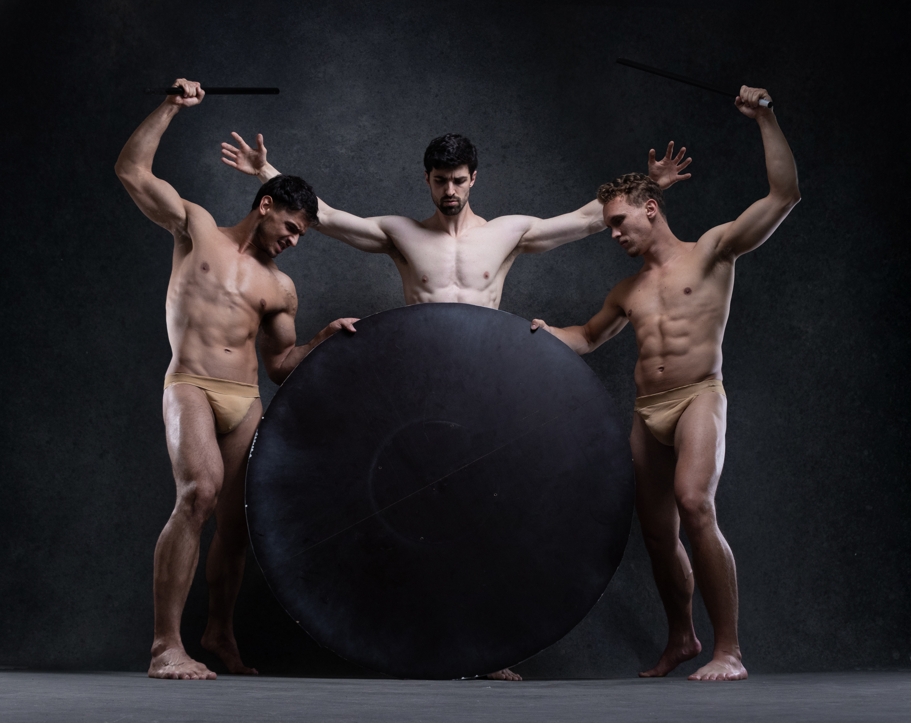 three muscular men in beige thongs pose with their bodies against a black background. the man in the center stands with his arms outstretched, while the men on either side of him stand with their arms raised above their heads, each holding a black stick. in the center of the image is a large black circle.