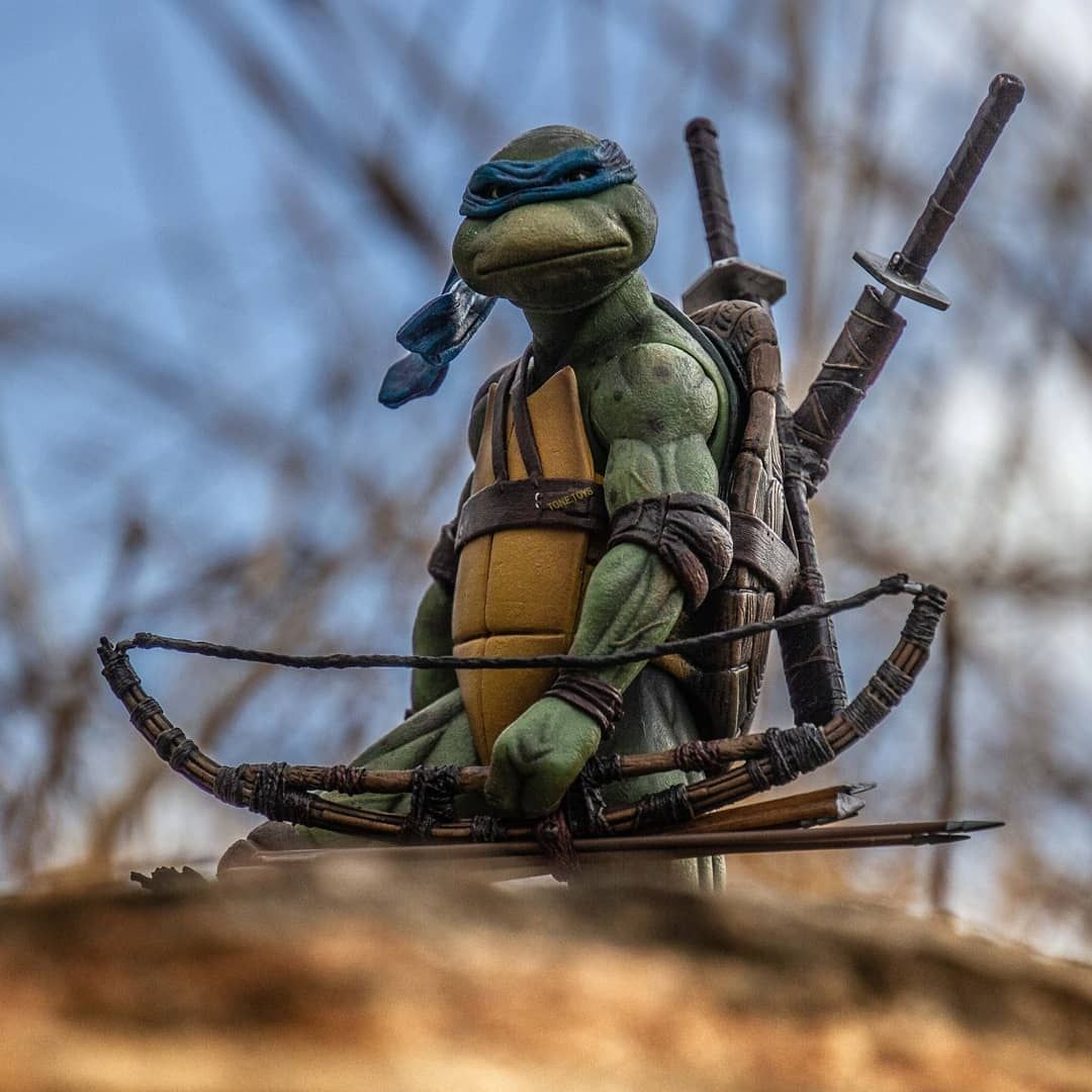 a closeup of a leonardo action figure from the teenage mutant ninja turtles, posed outdoors with a bow and arrow in front of him and his signature katanas strapped to his back. the figure is sitting on a textured surface that suggests a natural outdoor setting, likely a log or rock. the background is a blur of sky and trees.