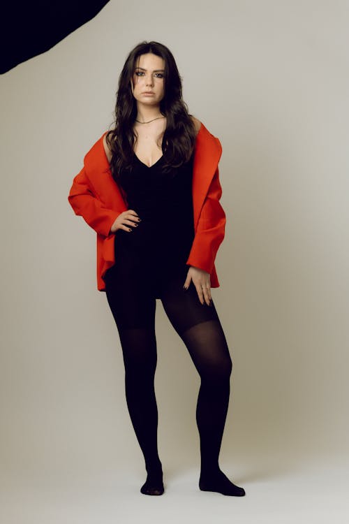 a full body studio shot of a woman with brunette hair wearing a black dress, black tights, and a red blazer. she is standing with her hand on her hip, looking at the camera. she is wearing a choker necklace. the background is a light gray color.