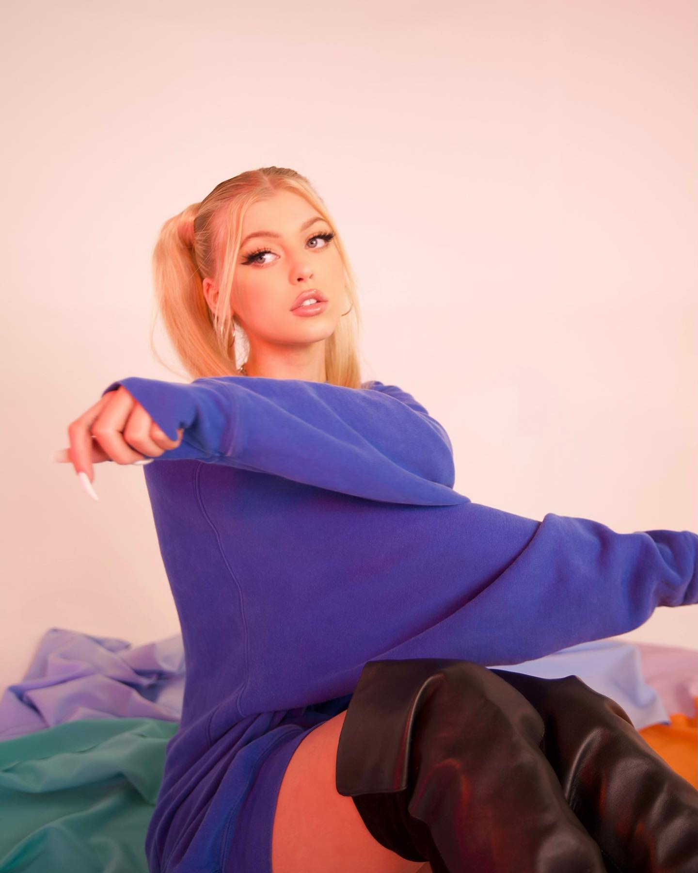 the image features a young woman with blond hair styled in pigtails, wearing a loose blue sweatshirt and black thighhigh boots. she is sitting on a bed with colorful bedding, posing for the camera with a direct gaze. her makeup is prominent, and she is wearing hoop earrings. the background is a plain, light color, likely in a studio setting.