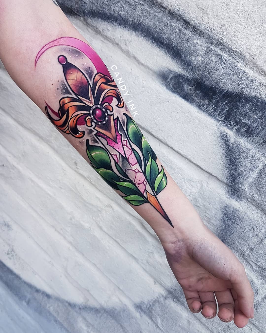 a closeup photo of a neotraditional style tattoo of a dagger on a forearm. the dagger has a crescent moon behind it and is decorated with green leaves, flowers and a pink jewel. the tattoo is done in a illustrative style with bold lines and vibrant colors.