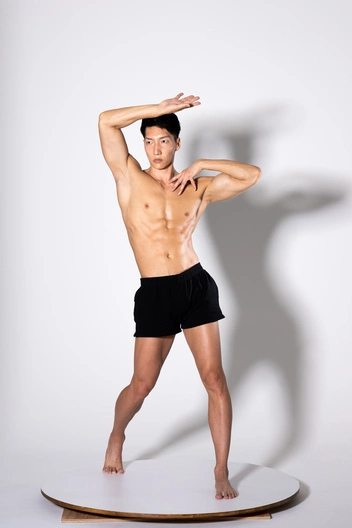 a photo of a muscular, shirtless asian man posing in black shorts on a round white platform against a white background.  he has his arms raised, one hand touching his forehead. his body is welldefined, showcasing a toned physique. the image is professionally lit, giving a clean and polished look.  the overall aesthetic is suggestive of fitness, dance, or modeling.
