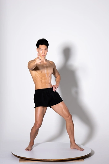 a muscular asian man stands on a white circular platform against a white background. he is shirtless, wearing black shorts, and has a confident expression. he is giving a thumbsup gesture with one hand, and his other hand is resting on his hip. his physique is lean and welldefined. the overall impression is one of health, fitness, and selfassurance.