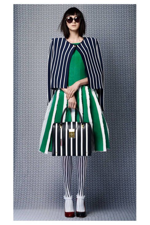 a fulllength studio shot showcases a fashion model elegantly dressed in a stylish ensemble. she's wearing a navy blue and white striped cropped cardigan over a vibrant green striped aline skirt. the skirt has a playful, slightly frayed hemline.  the model also sports black and white striped tights, black and white striped handbag with gold hardware, and black sunglasses. her shoes are black heels with a red accent.  the backdrop is a textured gray background with a subtle repeating pattern. the overall aesthetic is clean, modern, and sophisticated.