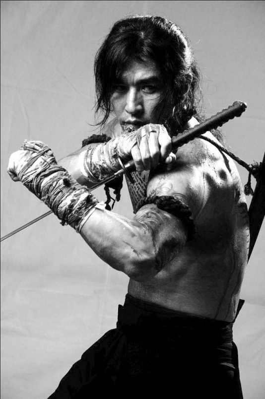 a muscular man in a black and white portrait is holding a katana, staring intently at the camera with a serious expression. he is barechested, wearing a black fabric belt and a long black fabric wrapped around his arm and wrist. the image is taken in a studio setting with a simple grey background. the man appears to be a  samurai or a warrior.