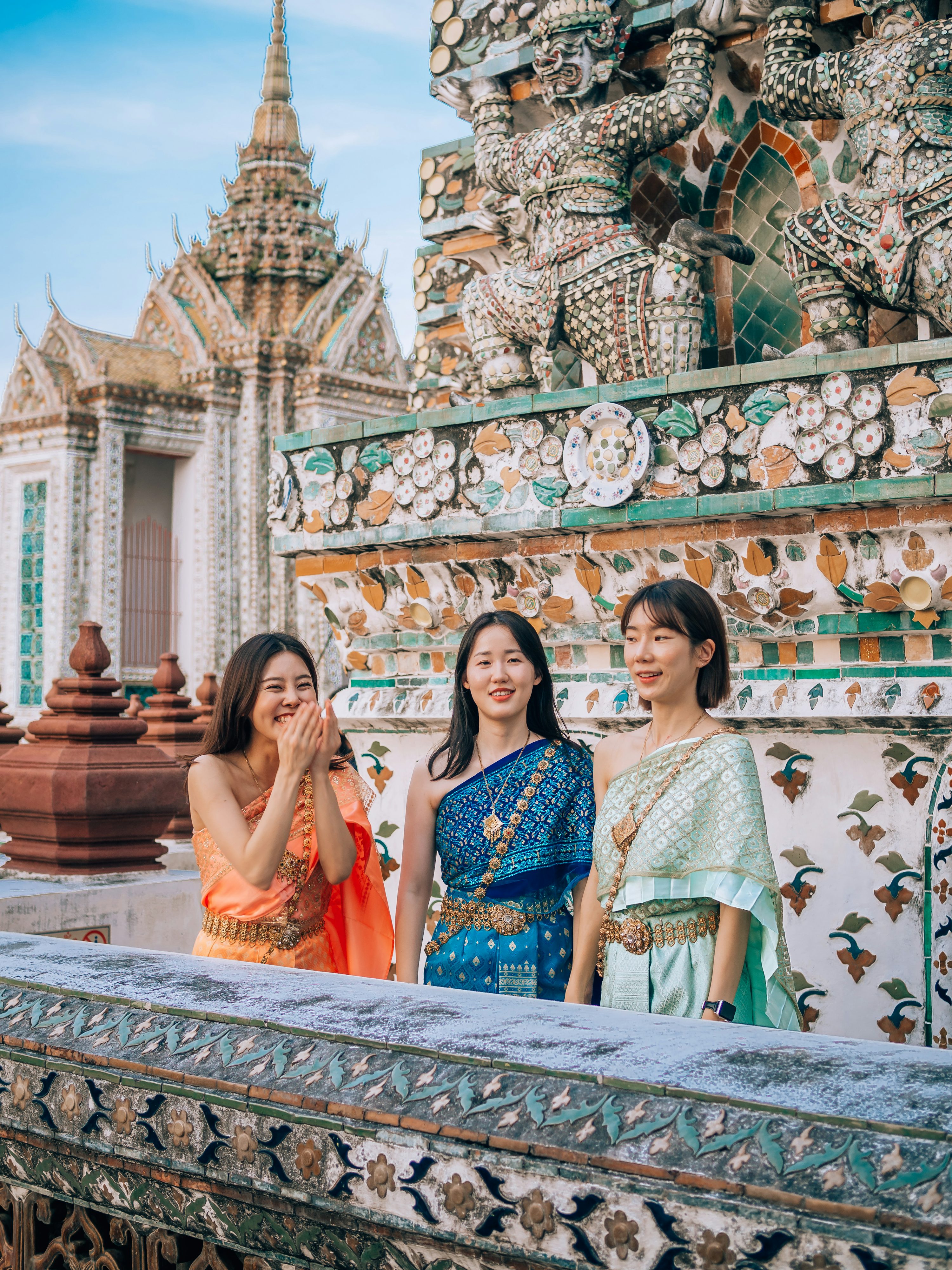 three young asian women, dressed in vibrant traditional thai clothing, stand smiling on a balustrade overlooking a section of wat arun ratchawararam ratchawaramahawihan (temple of dawn) in bangkok, thailand. the temple's intricate, colorful mosaics and statues are visible in the background. the women's attire contrasts beautifully with the rich textures and colors of the ancient temple setting. the scene conveys a sense of cultural immersion and joyful travel experience.