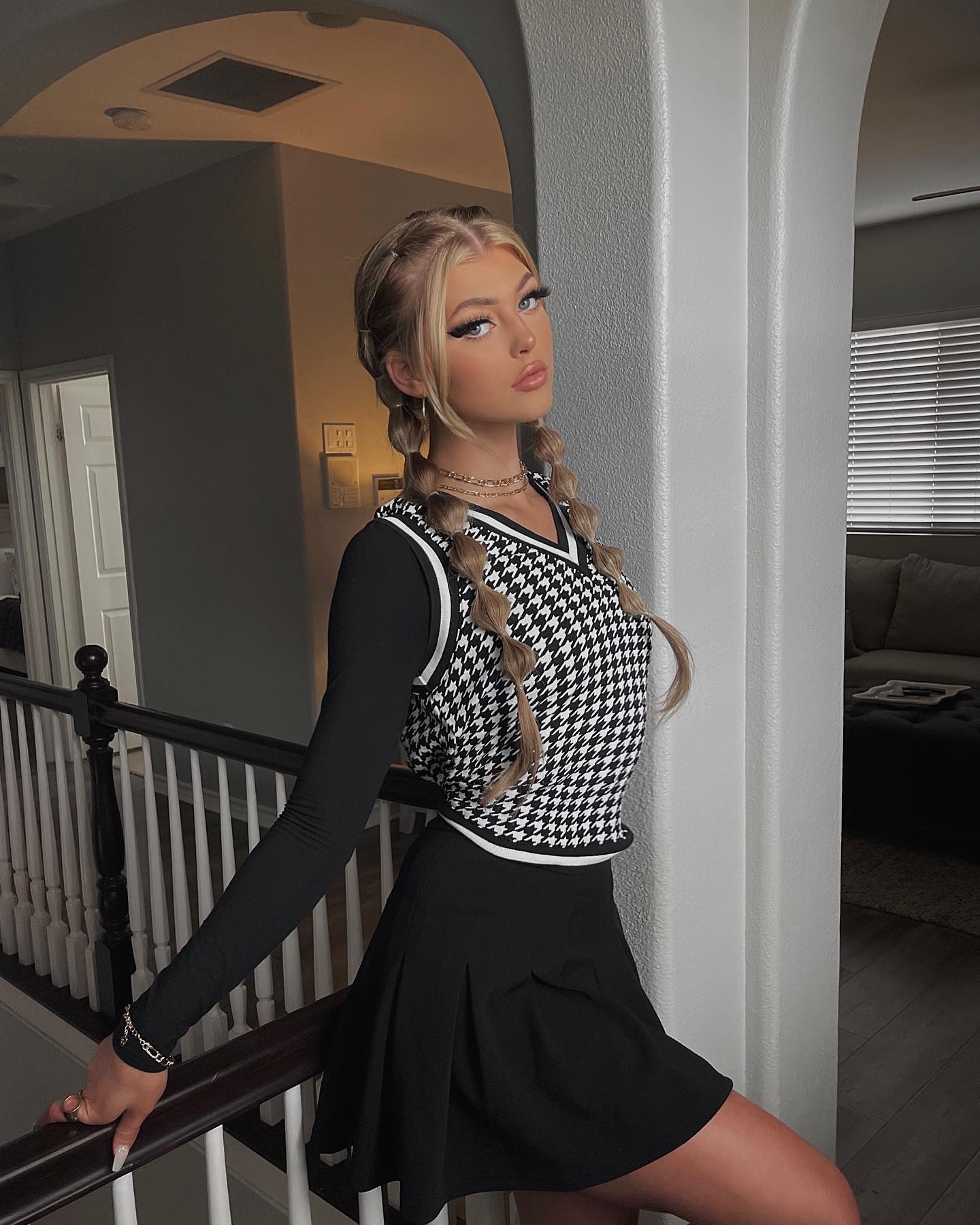 a young woman with blonde hair styled in braids stands in an indoor setting, leaning against a white column next to a staircase railing. she is wearing a longsleeved black shirt, a black and white houndstooth patterned vest, and a black pleated skirt. she is also wearing a necklace and a bracelet, and she is looking at the camera with a slight pout. the background includes a staircase, a doorway, and a glimpse of a living room with a couch and blinds on the window.
