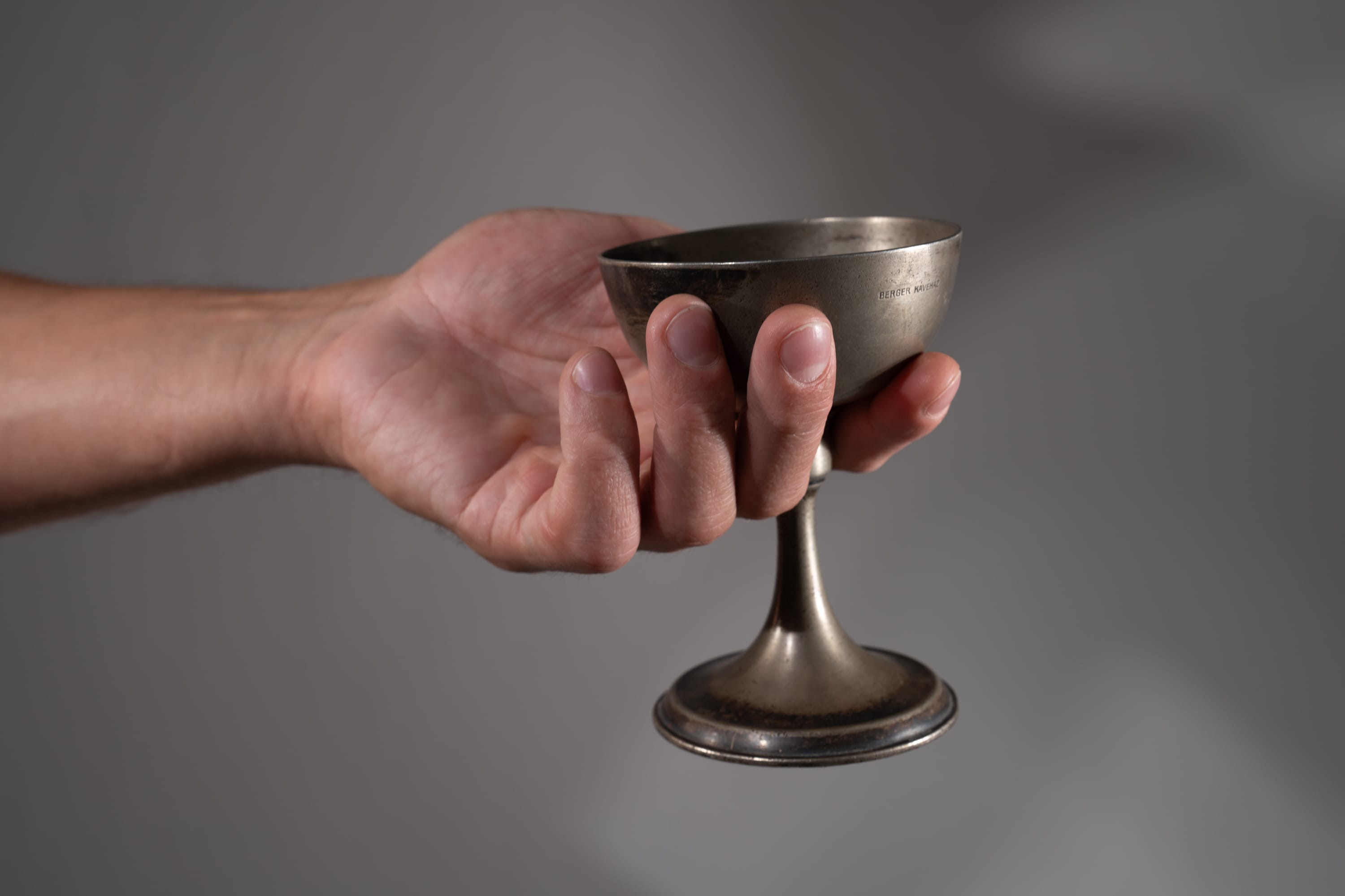 a closeup shot of a man's hand gently holding a tarnished, antique silver chalice.  the chalice is slightly tilted, as if ready to be filled. the background is a neutral gray, allowing the focus to remain on the object and the hand.