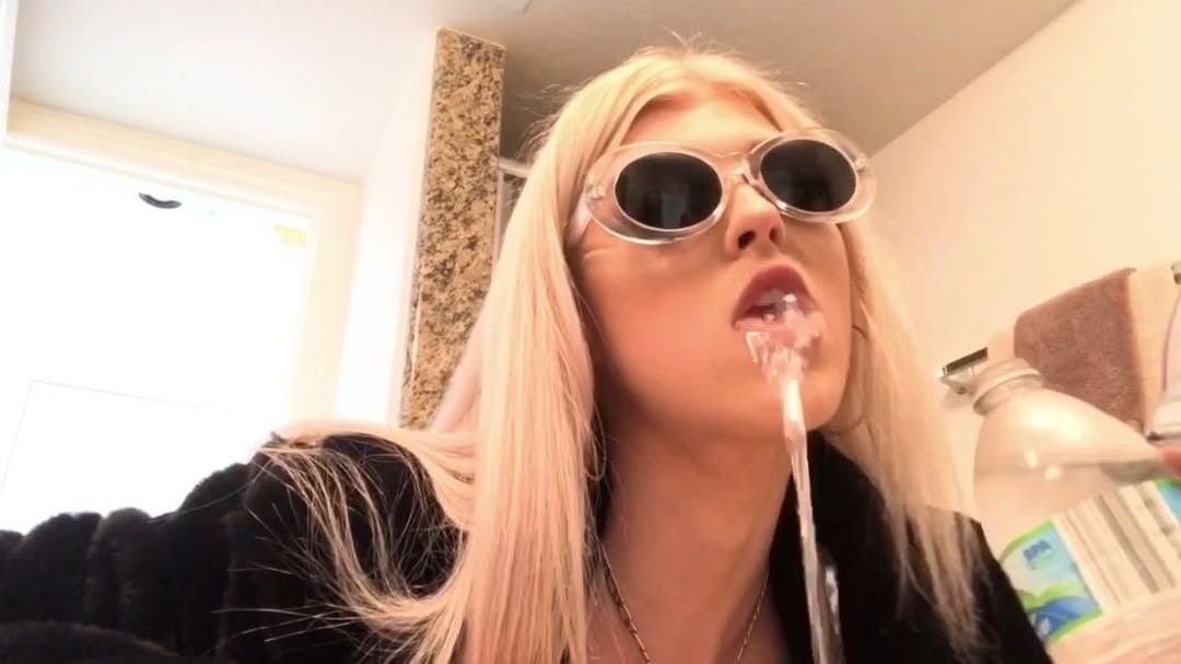 a woman with blond hair and sunglasses is drinking water from a bottle. she is indoors, possibly in a bathroom, and water is streaming from her mouth.