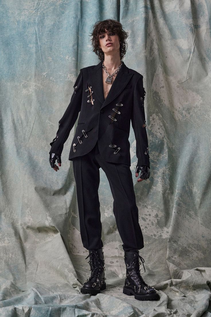 a young male model is posing in a dark, edgy outfit. he is wearing a black blazer with safety pins attached to the seams as embellishment, black pants, and chunky black combat boots adorned with more safety pins. the model also wears a silver necklace with a padlock pendant. the backdrop is a textured, light teal and gray fabric. the overall style is a blend of gothic and punk influences.
