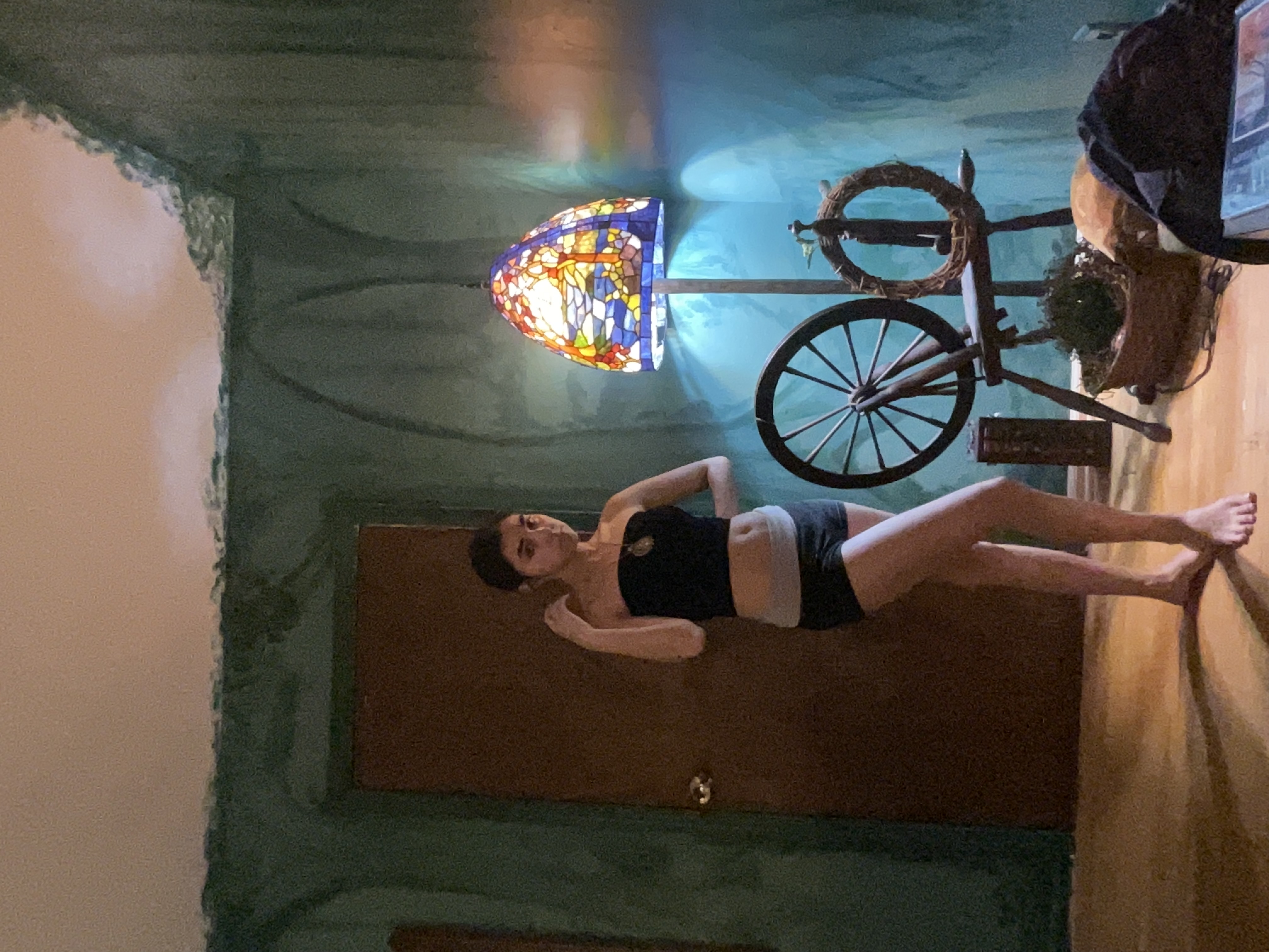 a young woman stands in a room with tealpainted walls. she is wearing a black crop top and grey and black shorts. there is a tiffanystyle stained glass lamp and a spinning wheel in the background. the room has a rustic, bohemian style. the woman is posed and looking at the camera.